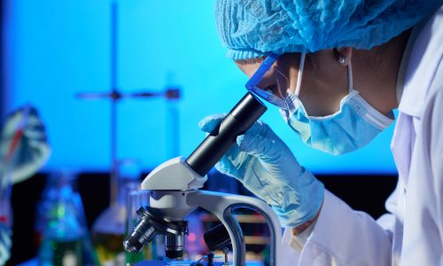 Profile view of concentrated Asian microbiologist examining sample with help of modern microscope while wrapped up in work at dim laboratory